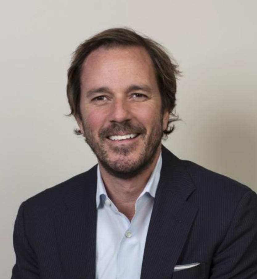 A smiling man with brown hair and a beard in a dark suit jacket and light blue shirt.