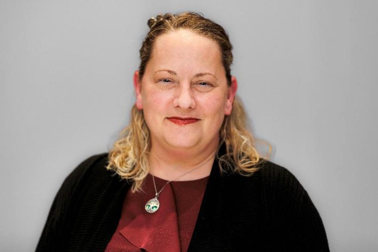 Headshot of a woman with curly blonde hair, wearing a red top, black cardigan, and a tree pendant.