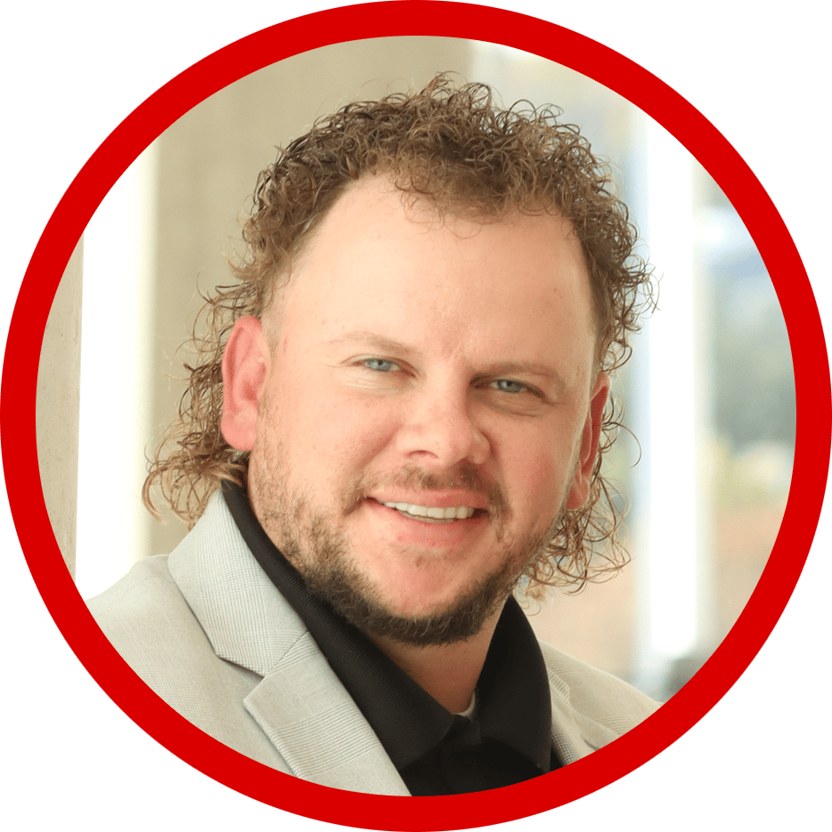 A smiling man with blue eyes, curly brown hair, and a beard, wearing a light gray jacket and black shirt.