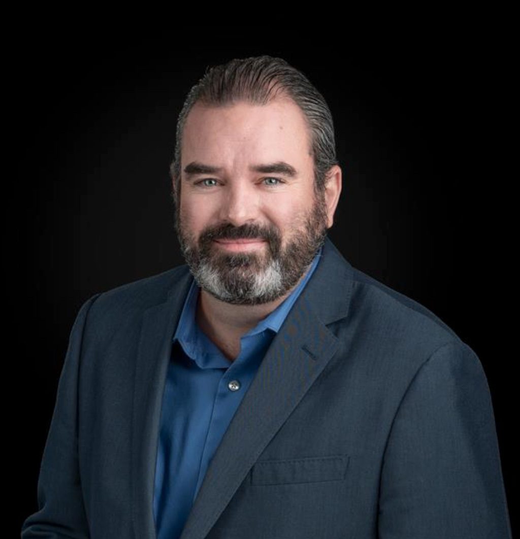 Headshot of a smiling man with a beard, blue shirt, and dark jacket against a black background.