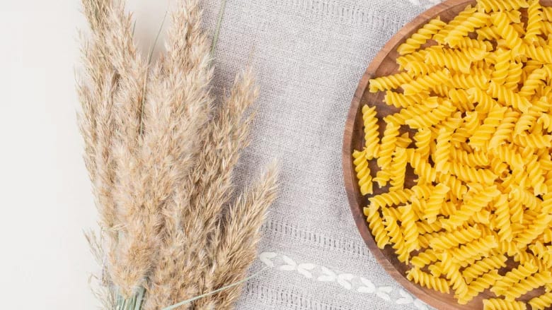 Dried pampas grass and uncooked fusilli pasta in a wooden bowl on a grey cloth.