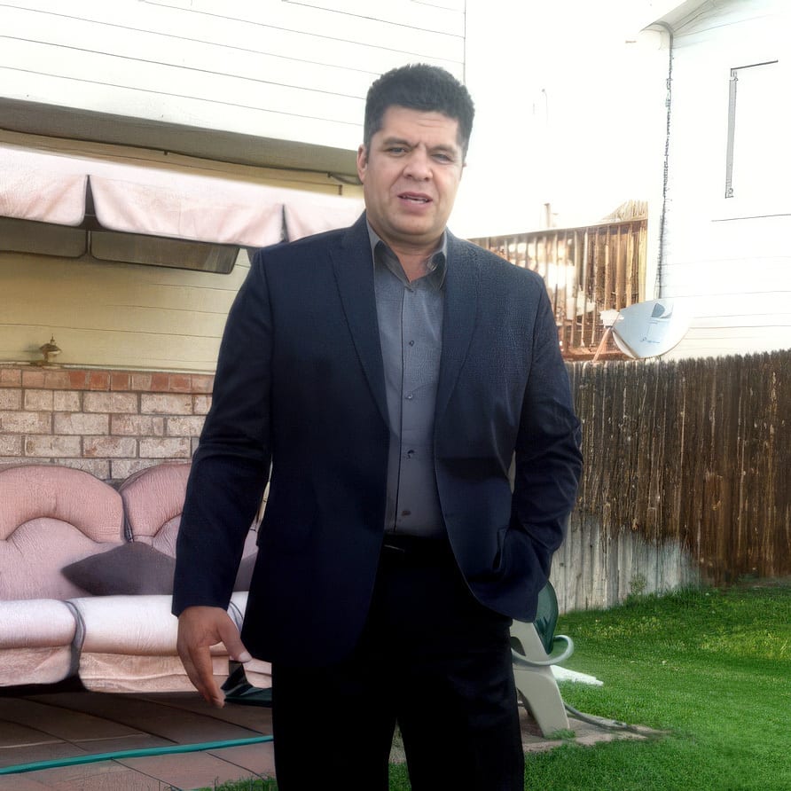 Man in a dark suit and grey shirt standing outdoors, looking at the camera.