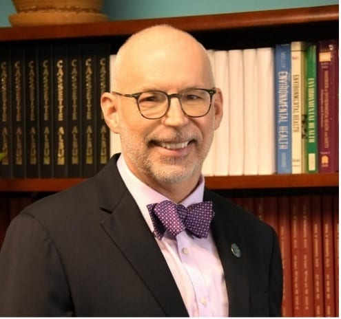 Smiling man with glasses and purple bow tie before "Environmental Health" books.