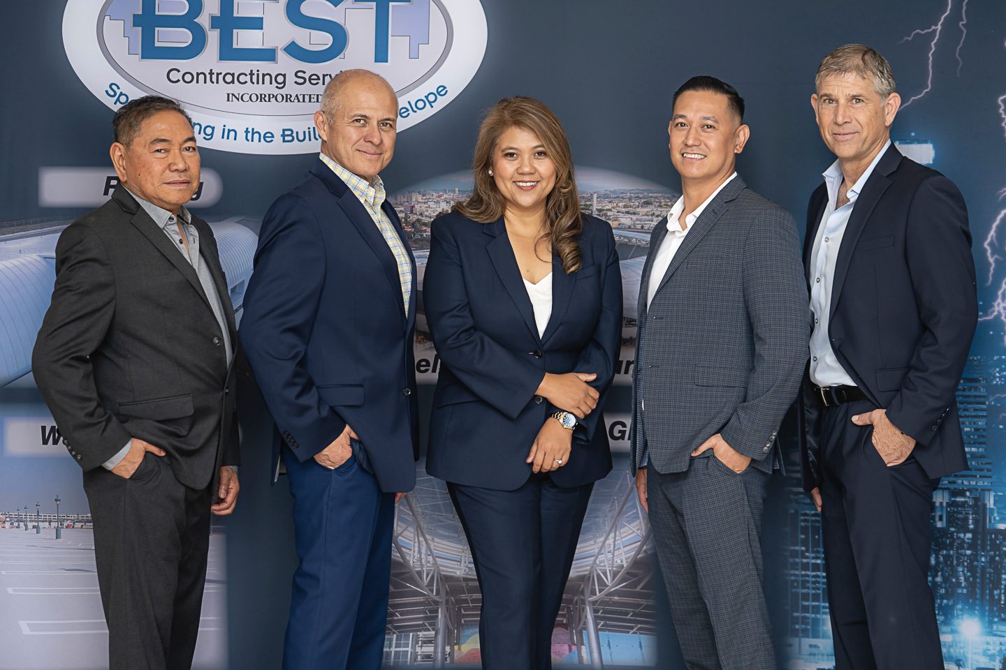 Five diverse professionals (4 men, 1 woman) in suits smiling against a company logo backdrop.