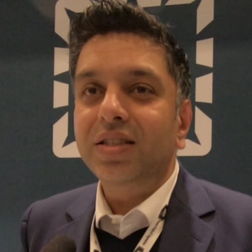 Man in suit talking, with a white angular logo in the background.
