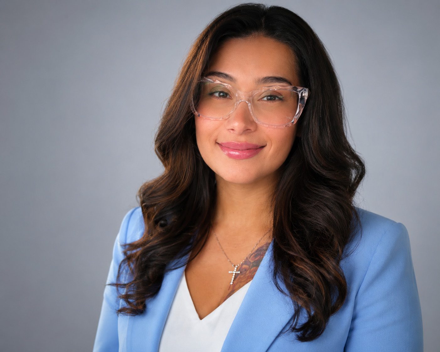 A smiling woman with dark wavy hair and clear cat-eye glasses wearing a blue blazer and white top.