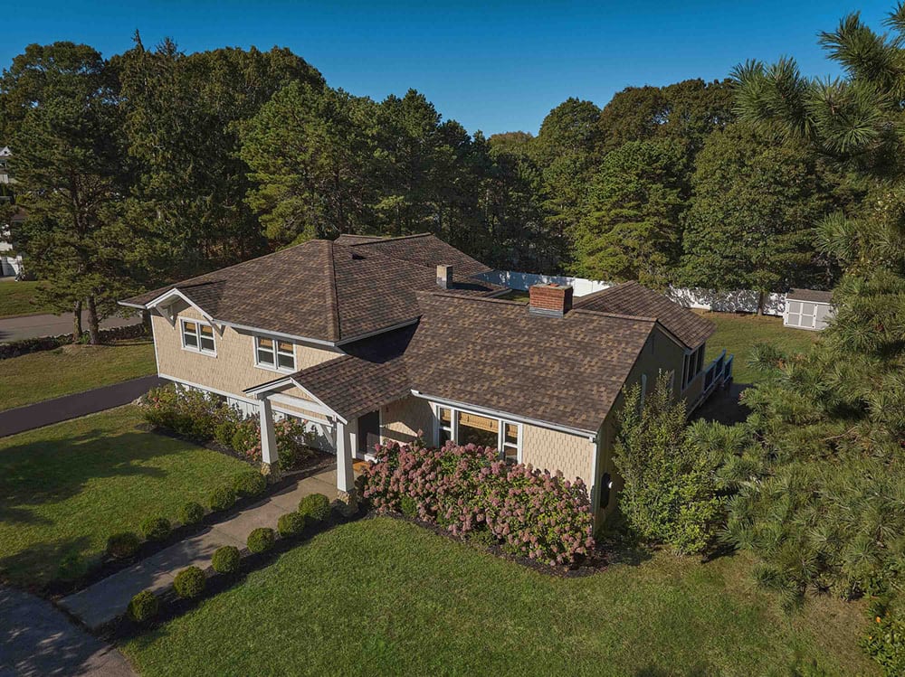 Aerial view of a house with a brown shingle roof, light siding, green lawn, and trees.