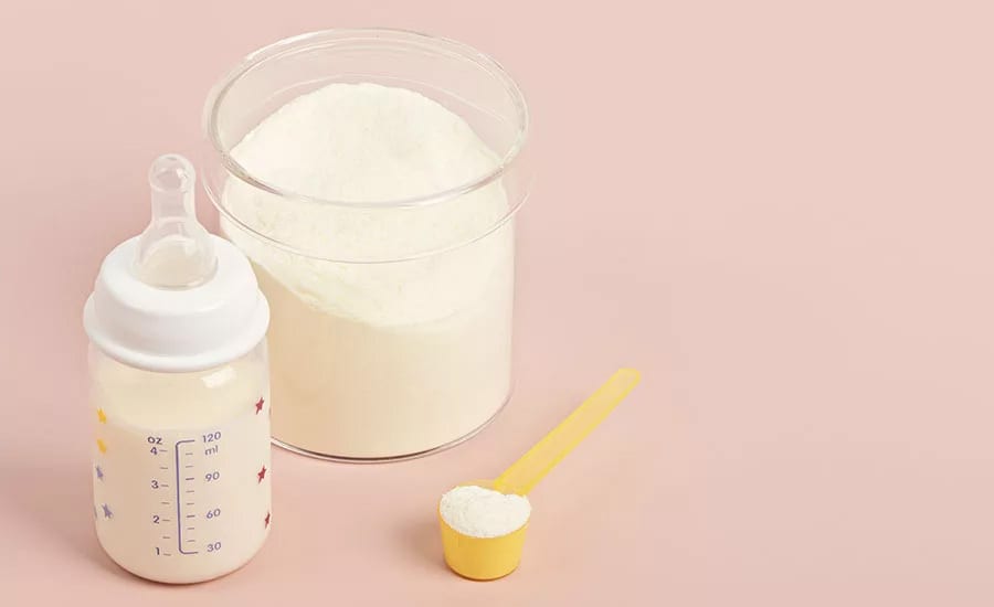 Baby bottle with milk, formula powder in a container and scoop on a pink background.