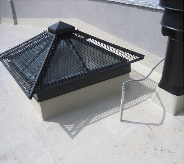 Skylight with black metal protective grate on a light-colored flat roof, next to a black pipe.