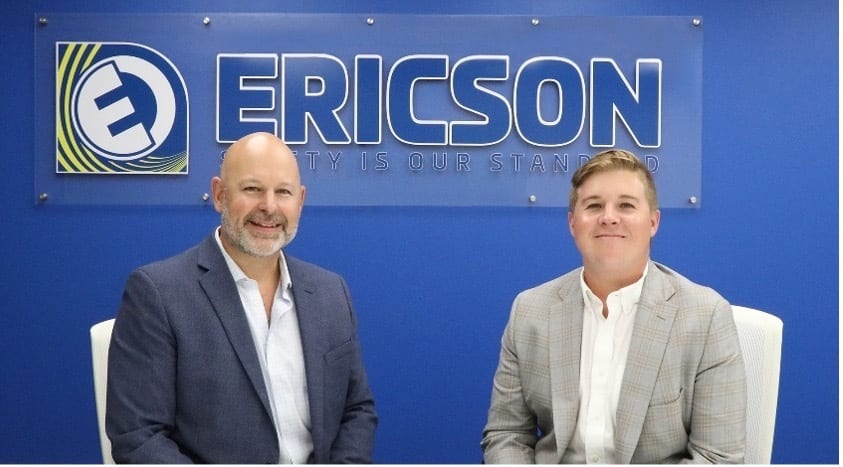 Two smiling men seated before an ERICSON sign on a blue wall.
