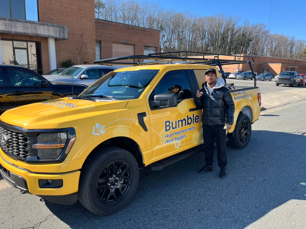 Man poses with a bright yellow pickup truck branded "Bumble Roofing" and a bee logo.
