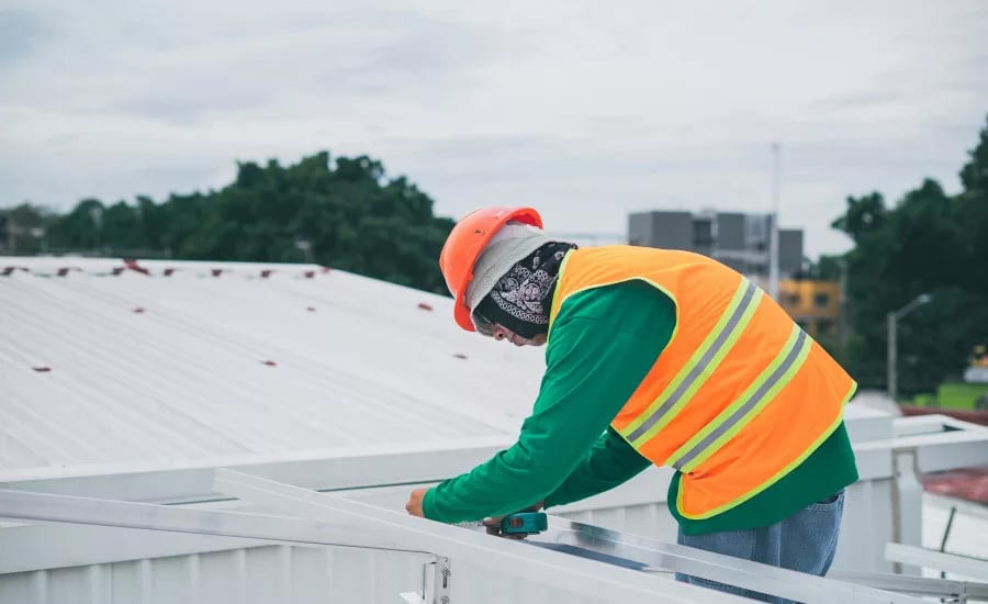Worker in hard hat and safety vest installing equipment on a white metal roof.
