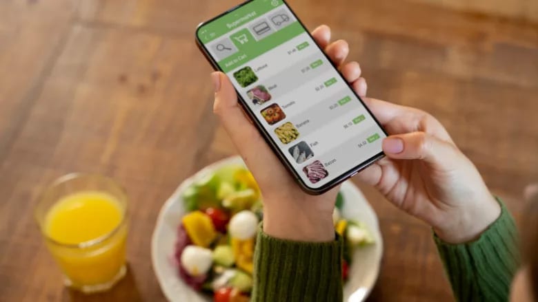 Person using a grocery shopping app on a smartphone, with a salad and orange juice on a wooden table.