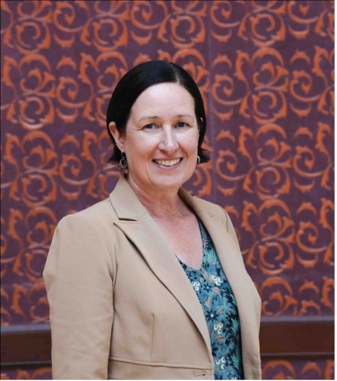 Smiling woman in beige blazer, patterned shirt, dark hair, against an ornate purple-orange wall.