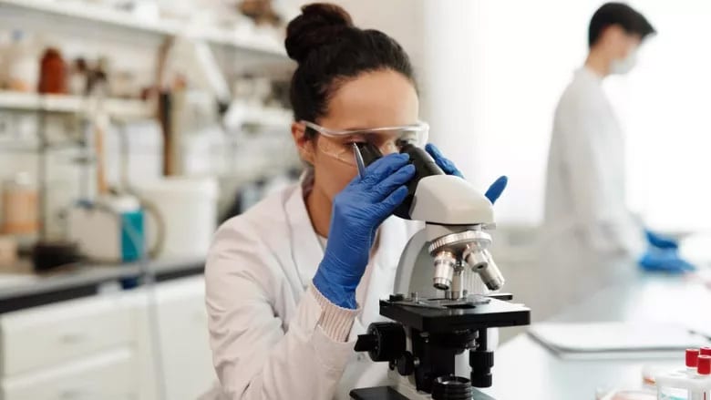 A female scientist in safety goggles and blue gloves looks into a microscope in a laboratory, with another person blurred in the background.