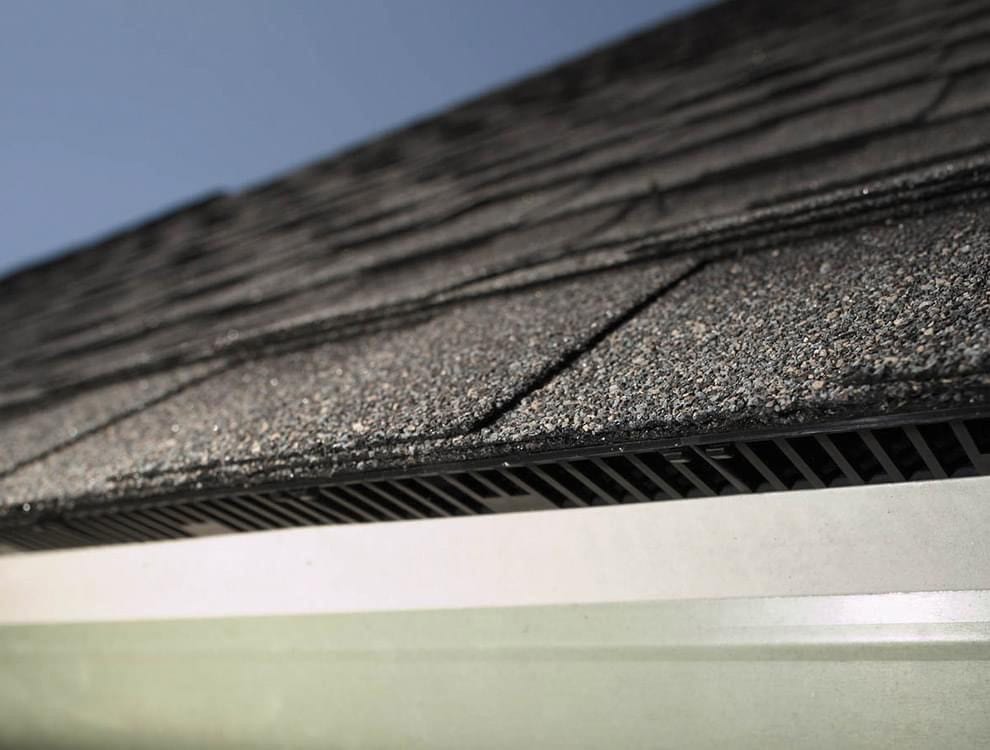 Black shingled roof edge with eave vent and white gutter.