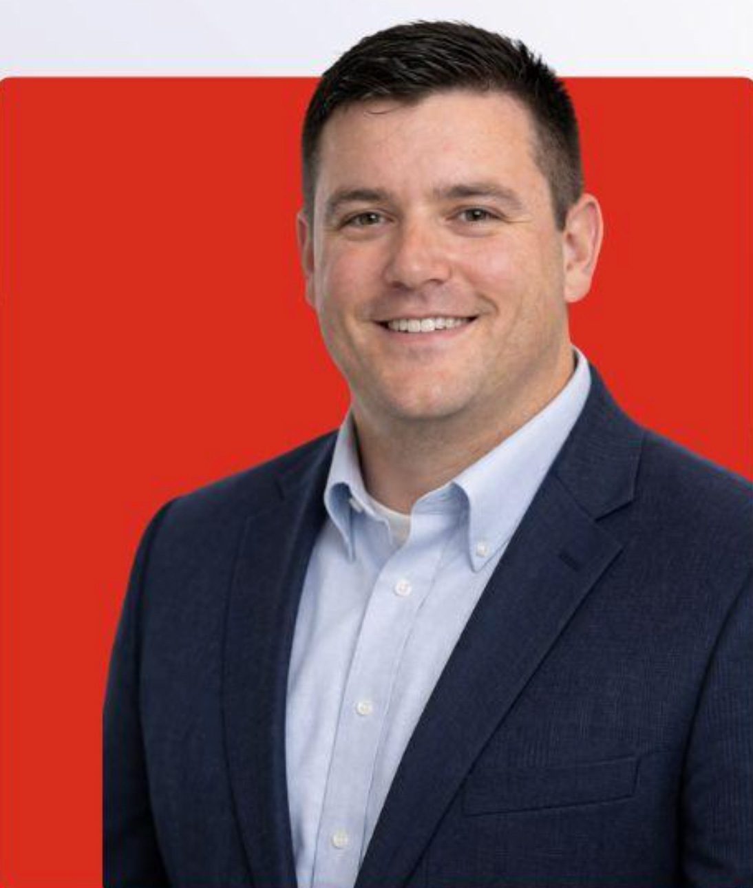 Headshot of a smiling man in a blue suit and light blue shirt against a red background.