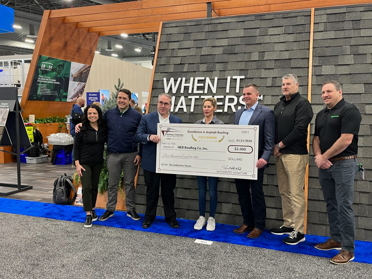 A group of seven people, including men and women, smiling and holding a large $2,000 check for HER Roofing Co. Inc.