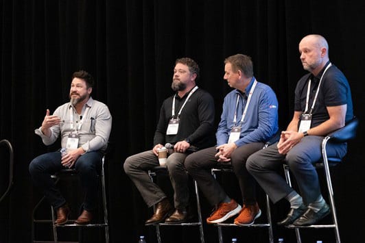 Four men on stage sitting on stools for a panel discussion; one gestures while speaking.