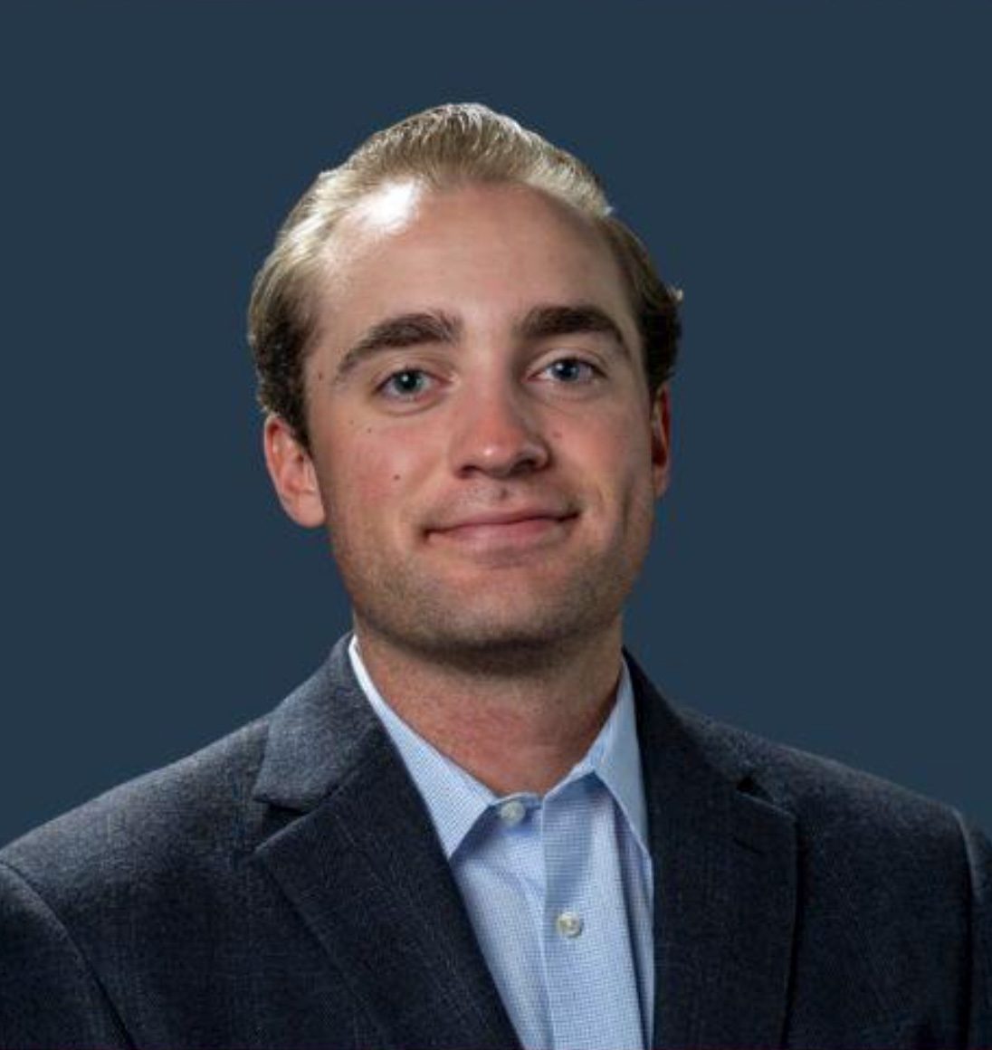 A young man with light hair and a light blue shirt under a dark suit jacket smiles.