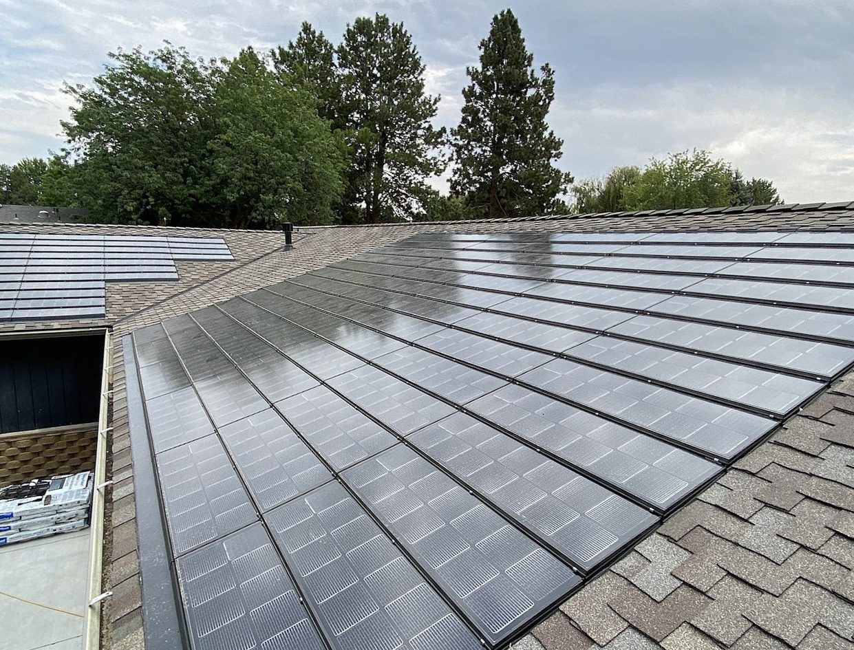 Integrated solar roof tiles cover a house roof, with asphalt shingles and trees under a cloudy sky.