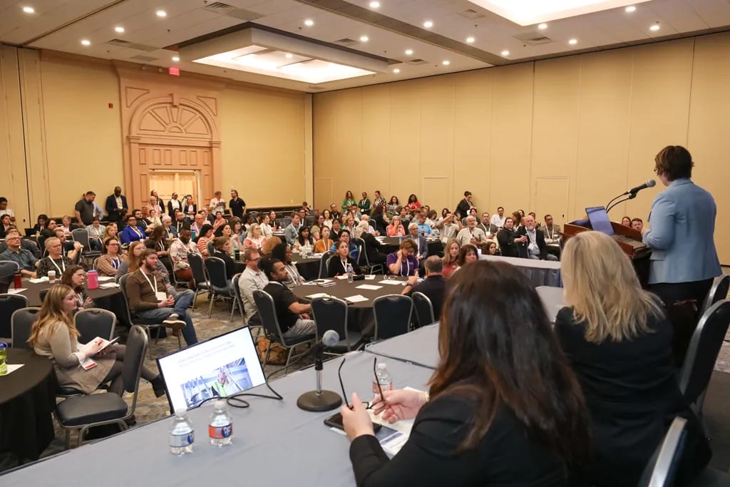 A speaker addresses a large audience in a conference room, with attendees at tables and a screen visible.