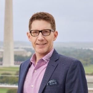 Smiling man in glasses and suit with Washington Monument in background.