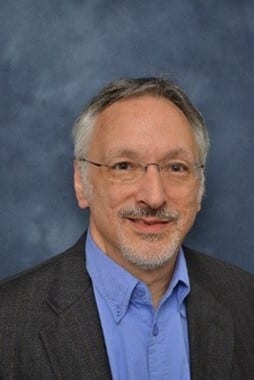 Man with grey hair, glasses, beard, blue shirt, dark jacket.