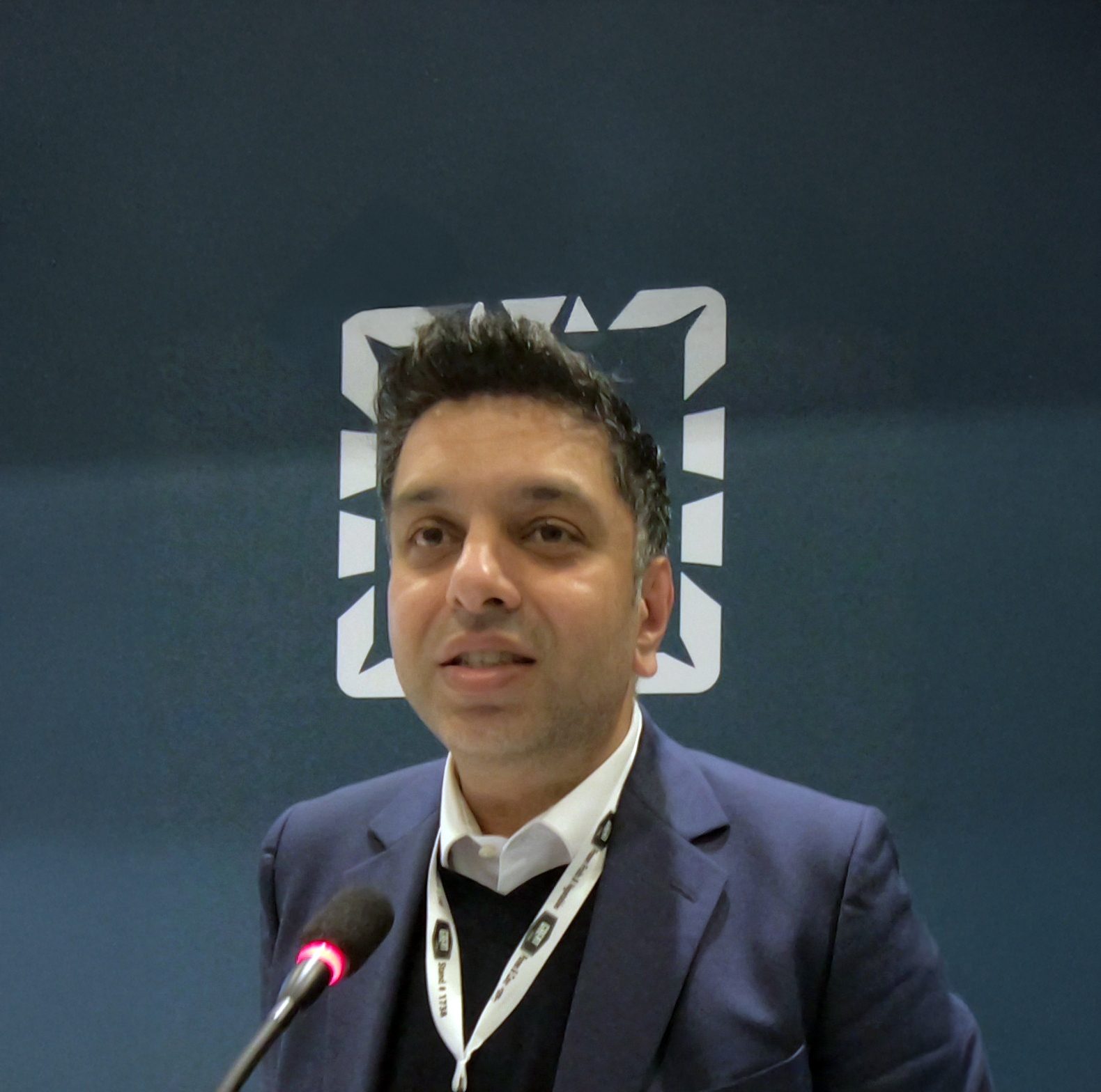 Man in a suit speaks into a microphone, with a white geometric logo on a dark background.