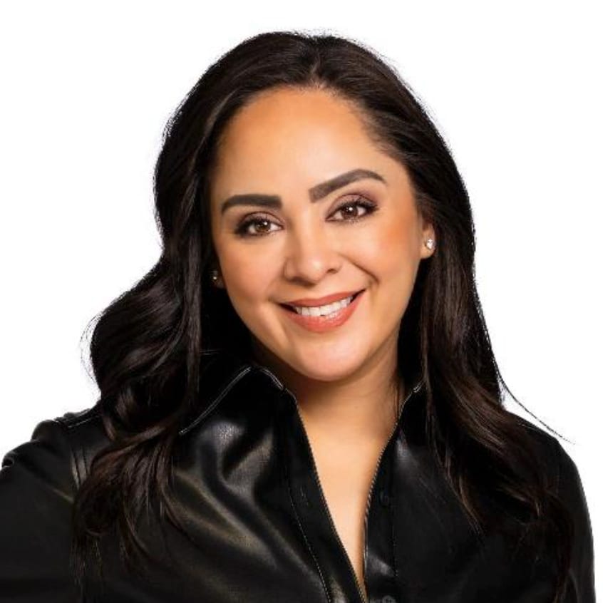 A smiling woman with long, dark wavy hair and a dark leather collared shirt, against a white background.