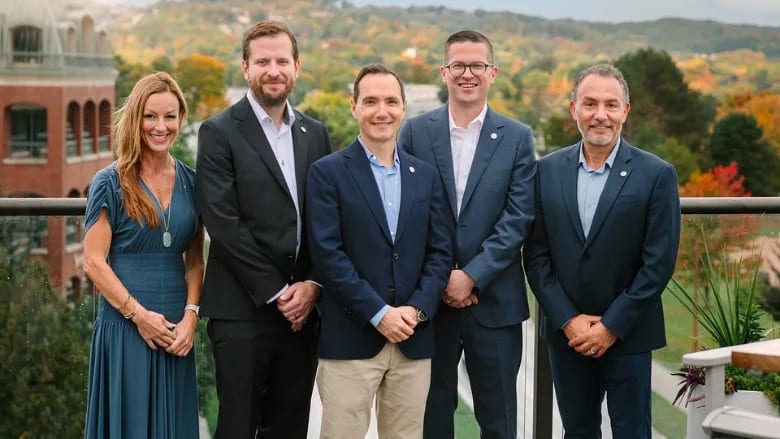 Five smiling people (1 woman, 4 men) in professional attire on a balcony with fall foliage.