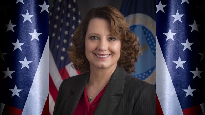 Smiling woman in a suit against American flags and a government seal.