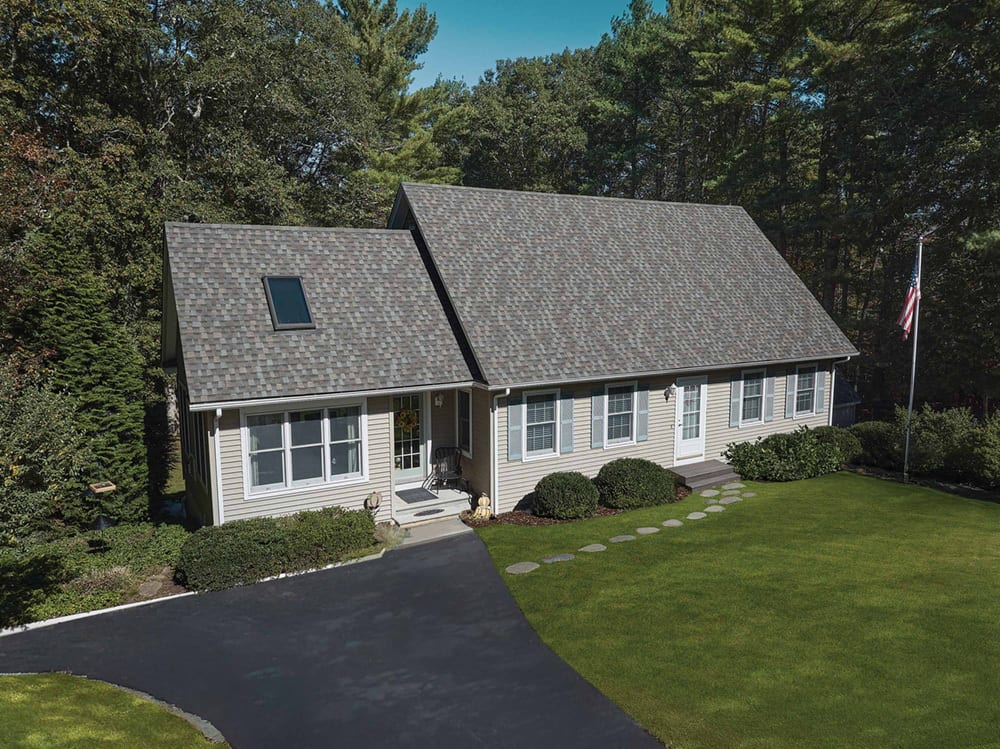 Aerial view of a house with gray roof, light siding, driveway, lawn, trees, and US flag.