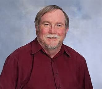 A smiling middle-aged man with a beard and mustache wearing a maroon collared shirt.