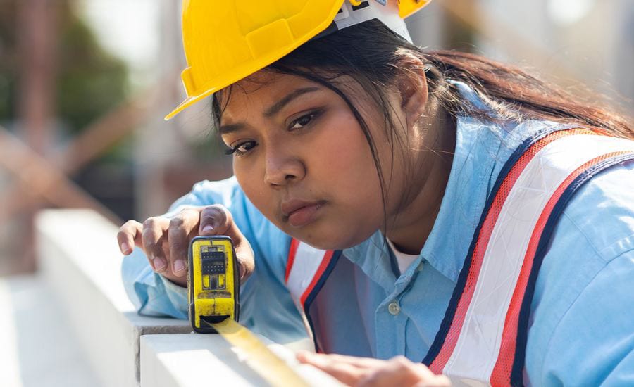 Person in hard hat and safety vest carefully measuring with a tape measure.
