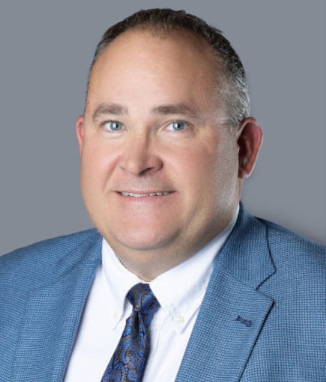 Smiling man in a blue suit and patterned tie, gray background.