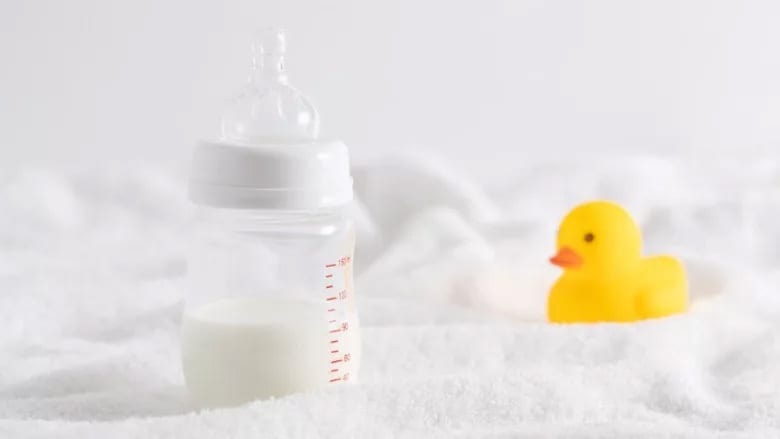 A baby bottle filled with milk next to a yellow rubber duck, both resting on a soft white surface.