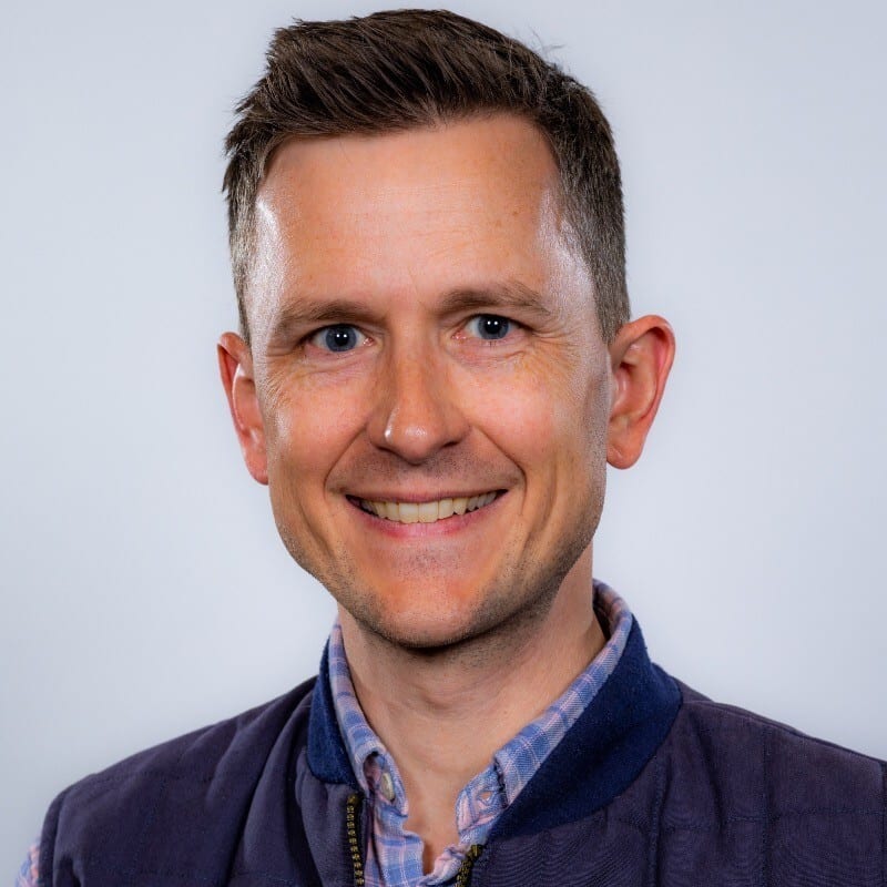 Smiling man with short brown hair, blue eyes, wearing a plaid shirt and navy vest.