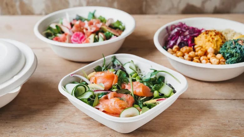 Three compostable bowls of diverse salads: salmon, chickpea, mixed greens, on a wooden table.
