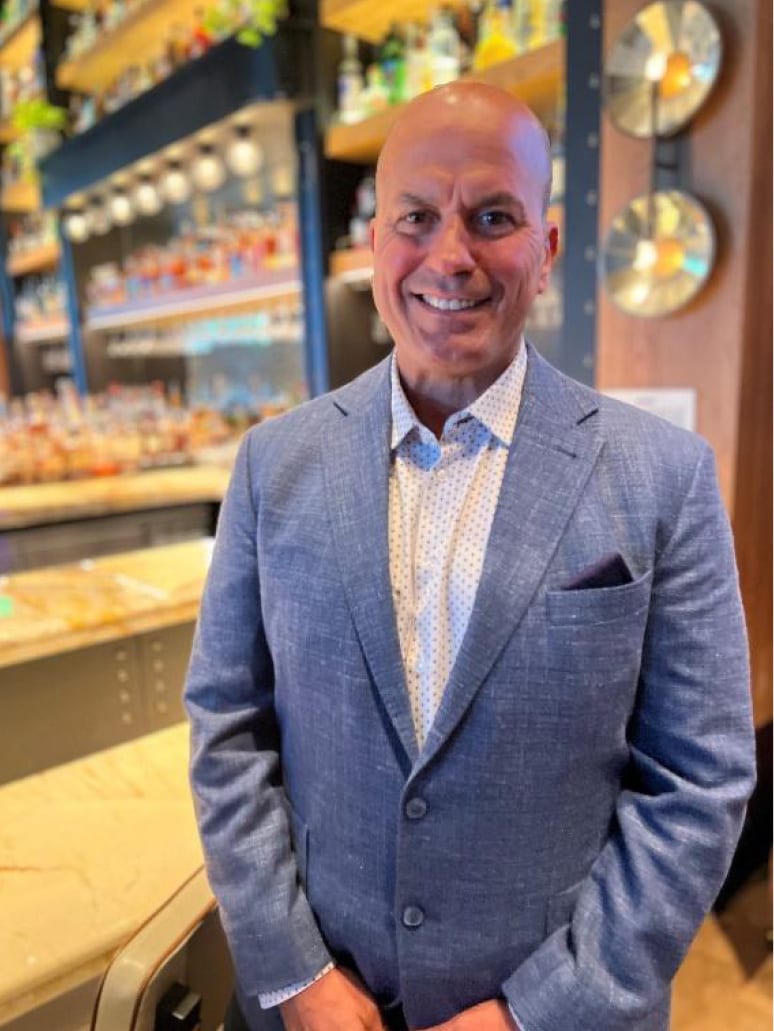 Smiling bald man in a blue suit jacket, standing in a bar with bottles in the background.
