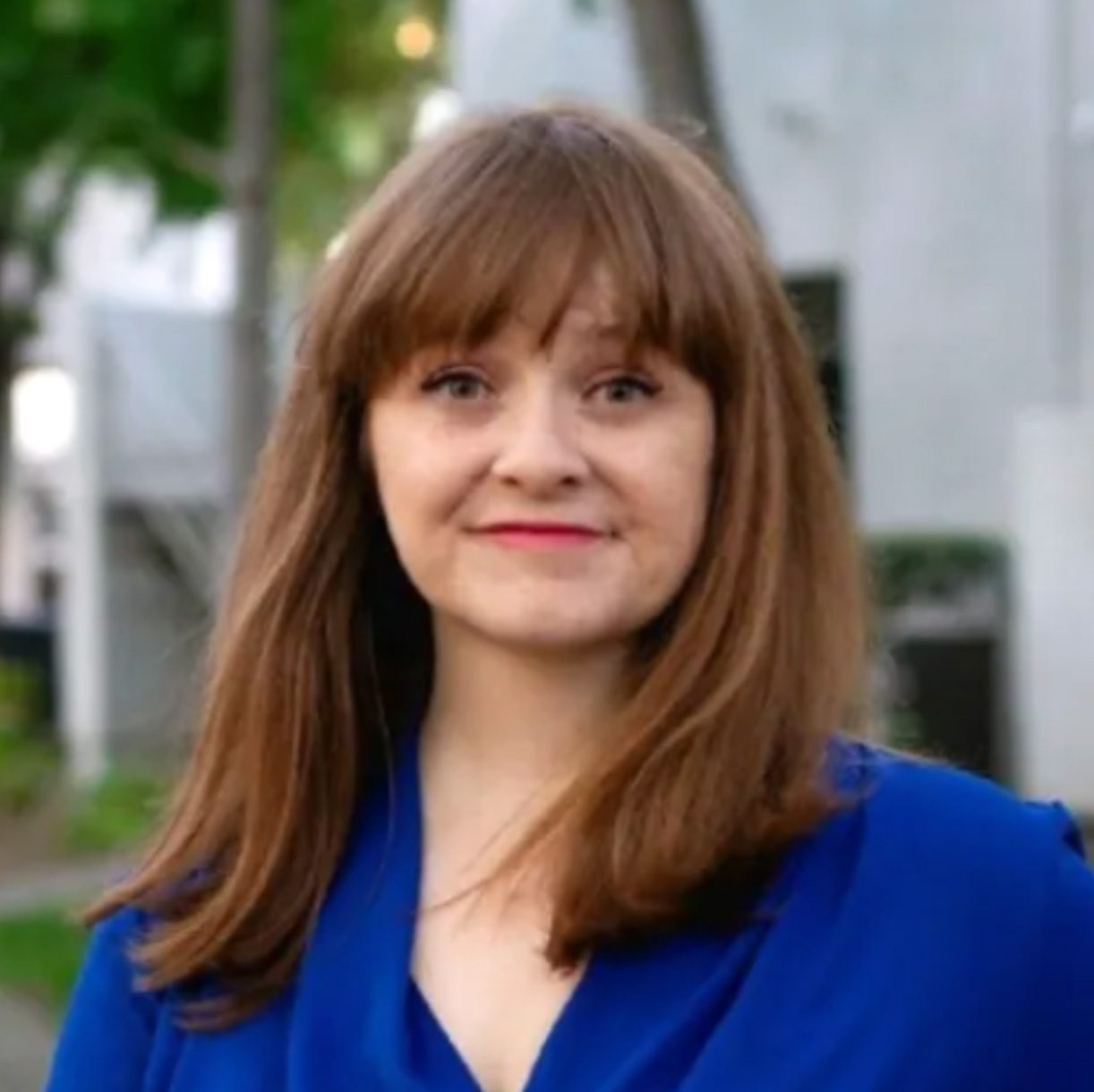 A smiling woman with brown hair and bangs, wearing a blue top, looks directly at the camera outdoors.
