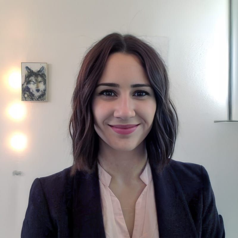 A smiling young woman with dark hair and a pink shirt under a dark blazer, looking directly at the viewer.