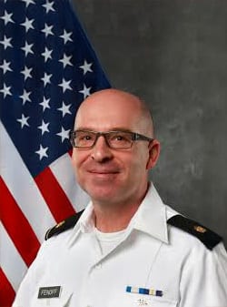 Smiling man in military uniform and glasses against an American flag.