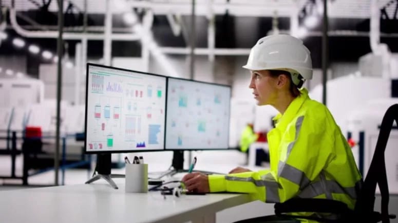 Engineer in hard hat & high-vis jacket monitors data screens in industrial setting.
