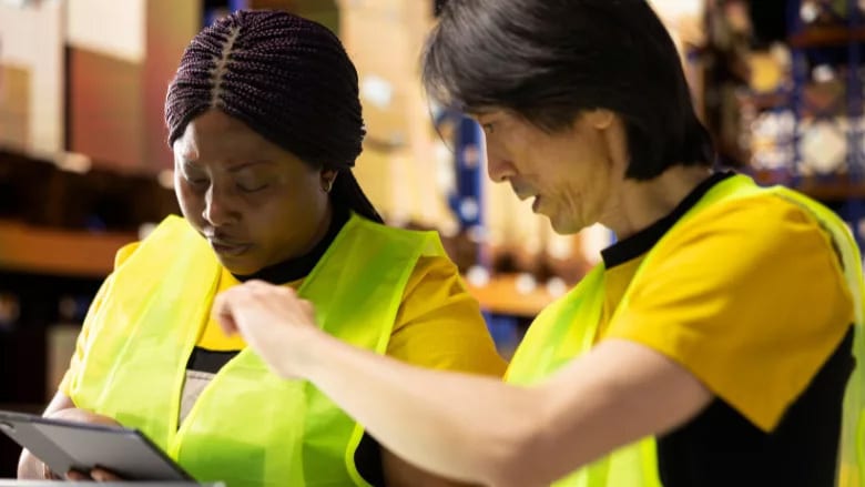 Two colleagues in safety vests checking data on a tablet in a warehouse.