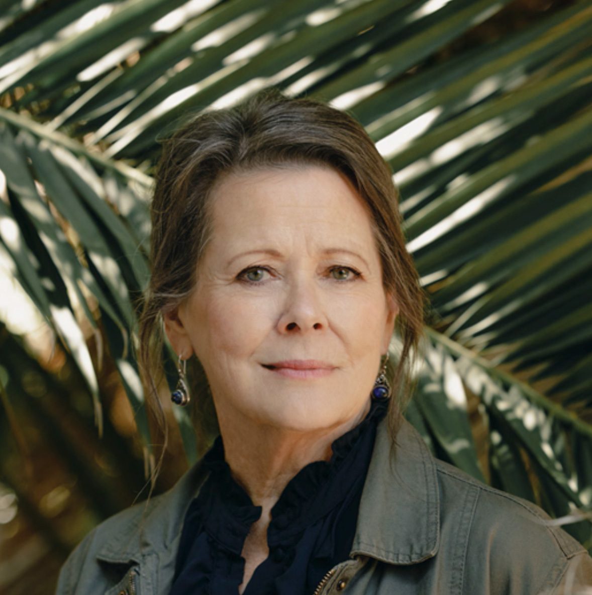Close-up portrait of a smiling woman with graying hair in an olive jacket against palm leaves.