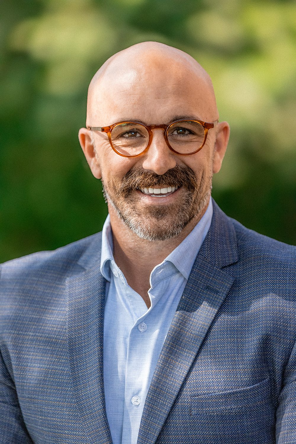 Smiling bald man with glasses and beard in a blue jacket against a green background.