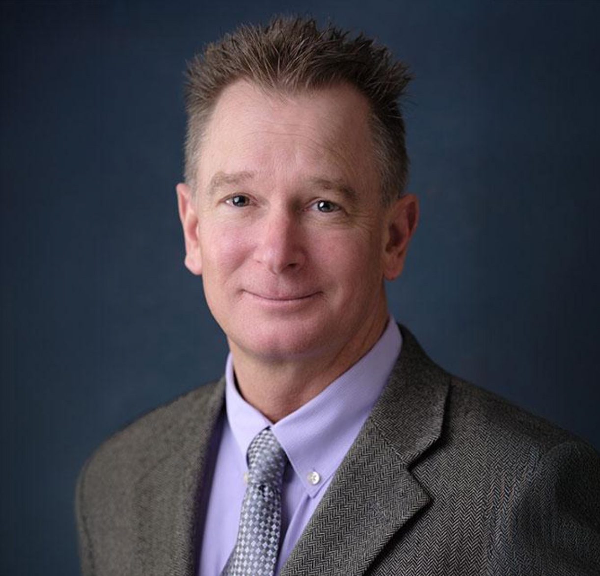 A smiling man in a purple shirt, gray tie, and suit jacket against a dark blue background.