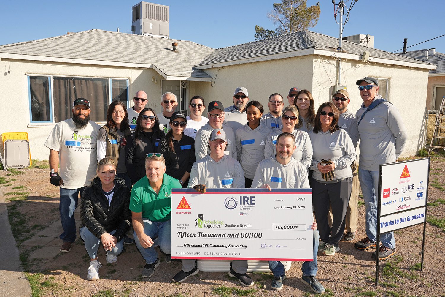 A group of volunteers stands outside a house, holding a large check for $15,000 for community service.