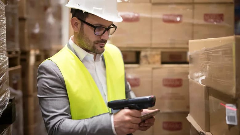 Man in hard hat and high-vis vest scanning boxes in a warehouse for inventory management.